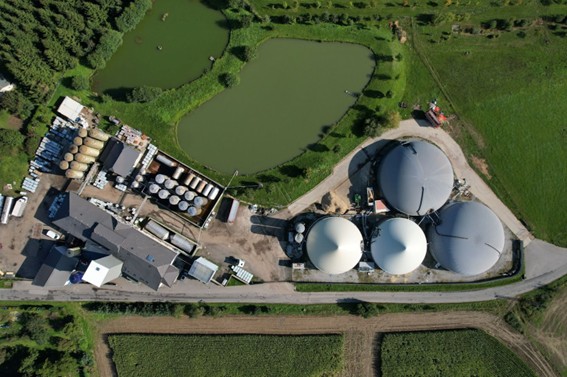Imagen aérea de una planta de biogás con tanques redondos, zona operativa y laguna artificial, rodeada de campos verdes y caminos rurales.