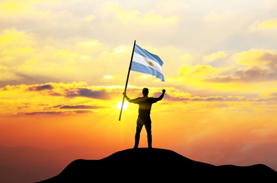 Persona de pie en la cima de una colina sosteniendo la bandera argentina con el sol poniente de fondo.