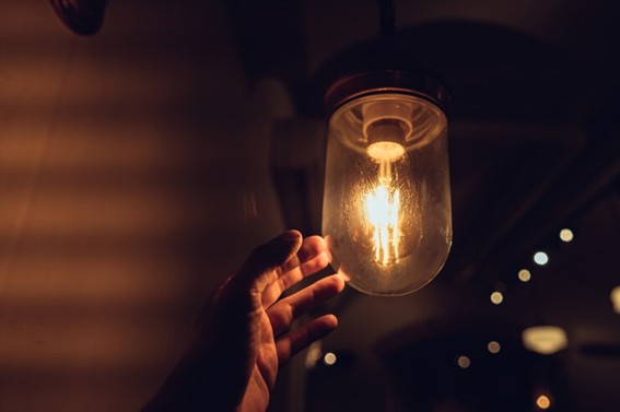 Mano humana acercándose a una lámpara encendida de vidrio en un ambiente oscuro y cálido, con luces suaves de fondo.