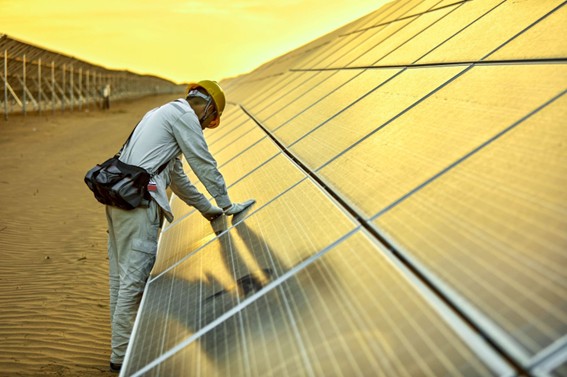 Trabajador con casco inspecciona paneles solares en un parque ubicado en el desierto, con luz cálida del atardecer reflejada en las superficies.