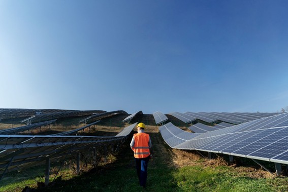Persona con casco y chaleco de seguridad caminando entre paneles solares en un campo, bajo un cielo azul despejado, en una planta de energía fotovoltaica.