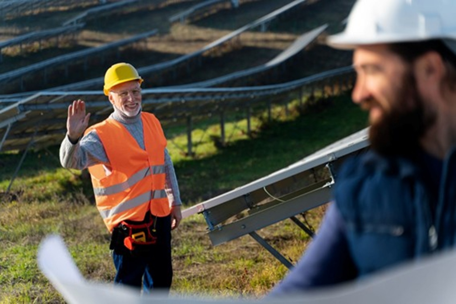 Trabajador con chaleco naranja y casco amarillo saluda a un compañero con casco blanco mientras supervisan paneles solares en campo.