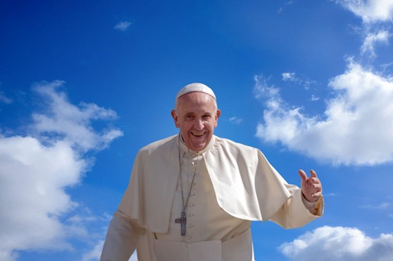 Persona vestida con sotana blanca y cruz al cuello levantando la mano en gesto de saludo bajo un cielo azul con nubes.